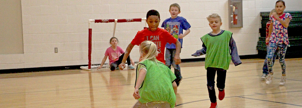A picture of multiple young students in a gym playing soccer.