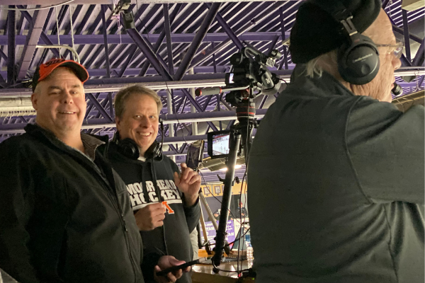 In 2019, Gregg Gess and Mark Moilanen, boys hockey parent helpers, stream a game in Buffalo with Larry announcing. 